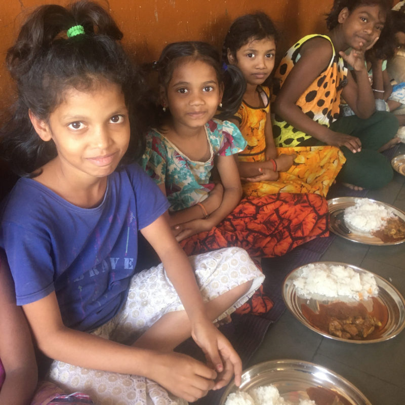 Four child residents at Rainbow Homes smile at camera whilst sitting down to eat lunch.